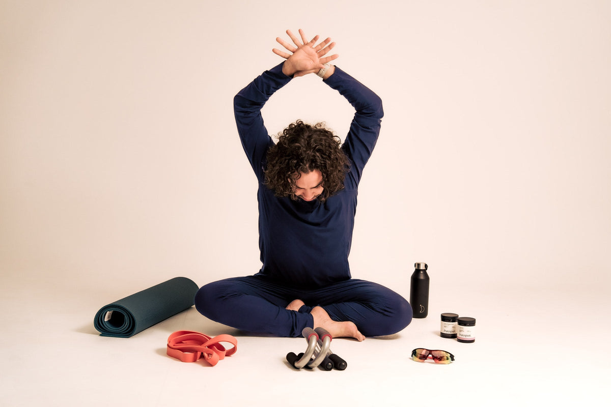Man in navy Zed long-sleeve set seated cross-legged with arms stretched overhead, surrounded by a yoga mat, resistance bands, push-up handles, water bottle and supplements