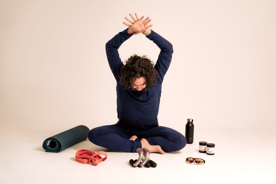 Man in navy Zed long-sleeve set seated cross-legged with arms stretched overhead, surrounded by a yoga mat, resistance bands, push-up handles, water bottle and supplements