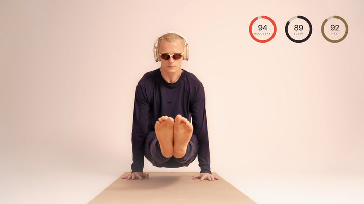 Man in navy ZedCore long-sleeve top and pants seated on a yoga mat with headphones, with three small recovery score rings overlaid in the corner