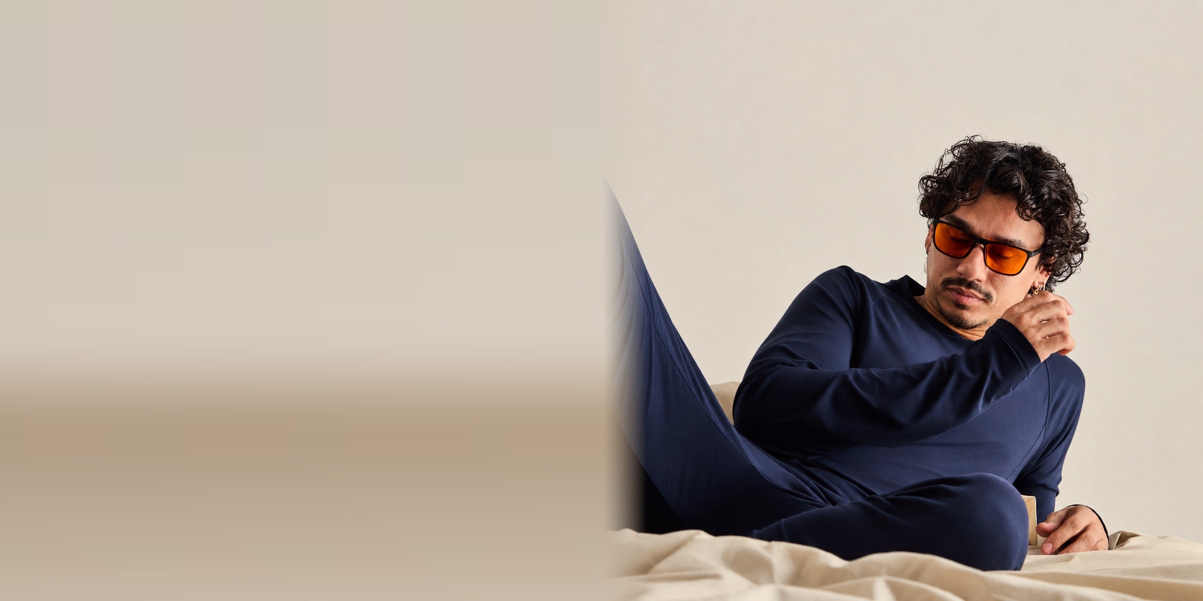 Man reclining on cream bedding in a navy Zed long-sleeve set wearing blue-light glasses