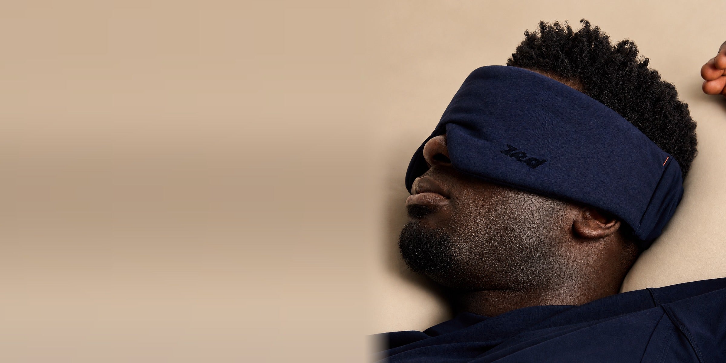 Man lying on cream bedding wearing the navy Zed sleep mask